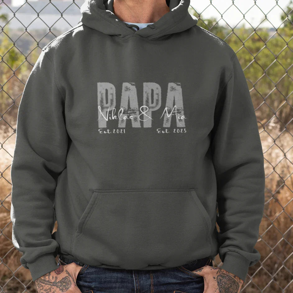 Dunkelgrauer Pullover Hoodie mit 'DADA Niklas & Mia Est 2021 Est 2023' Aufdruck, tätowierter Mann